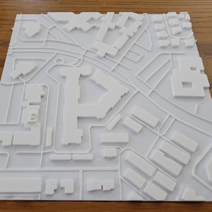 May include: A white 3D printed model of a city with buildings and streets. The model is made of plastic and is about 10 inches wide.