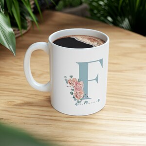 Può includere: Una tazza in ceramica bianca con una lettera F blu e un design floreale. La tazza è piena di caffè.