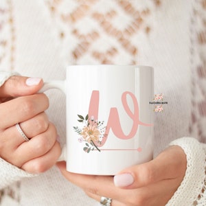 May include: White ceramic mug featuring a pink floral design and the letter "W" in pink. The mug is being held by a hand.