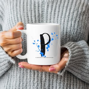 Può includere: Tazza in ceramica bianca con la lettera "P" nera e schizzi blu. Una linea tratteggiata è sotto la lettera. La tazza ha un manico. La persona indossa un maglione grigio.