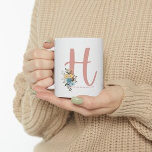 Puede incluir: Taza de cerámica blanca con una gran letra "H" rosa polvoriento y detalles florales en azul, amarillo y verde. Una línea de puntos está debajo de la letra. La taza está sujeta por una persona.