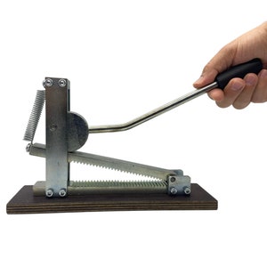 Op de afbeelding: Een zilveren handbediende gereedschap met een zwarte handgreep op een houten basis. Het gereedschap heeft een veer en een tandwielmechanisme.