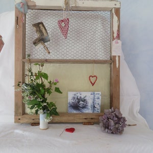 May include: A rustic wooden window frame with a wire mesh insert, decorated with red and white hearts, a small bouquet of flowers in a white vase, and a vintage postcard.