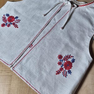 Könnte beinhalten: Eine weiße Weste mit roter und blauer Blumenstickerei. Die Weste hat einen roten Zickzackbesatz an den Kanten und einen Kordelverschluss am Halsausschnitt. Die Stickerei zeigt eine zentrale rote Blume mit blauen Akzenten.