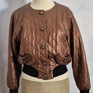 May include: A metallic bronze quilted bomber jacket with gold buttons and black ribbed cuffs and hem. The jacket is displayed on a mannequin with a cream turtleneck.