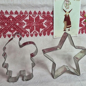 May include: Two metal cookie cutters: a star and a Santa Claus figure. The silver cutters are on a white surface with a red and white patterned border. A small illustration is in the background. Ideal for festive baking.