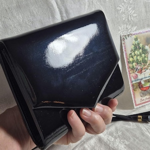 Könnte beinhalten: Eine glänzende, schwarze Clutch mit Klappenverschluss. Die Tasche wird in einer Hand gehalten und hat eine reflektierende Oberfläche. Eine Vintage-Weihnachtskarte mit einem Engel und einem geschmückten Baum befindet sich im Hintergrund. Zwei schwarze Riemen mit goldenen Akzenten sind ebenfalls sichtbar.