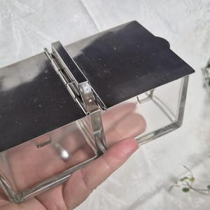 May include: A vintage glass and metal hinged box with a rectangular shape. The box has a clear glass body and a shiny silver metal lid and hinge. The box is held in a hand.