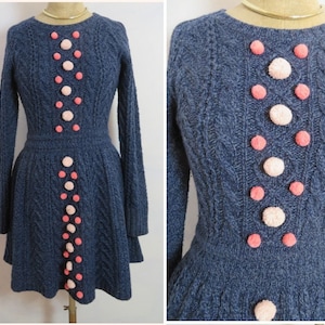 Könnte beinhalten: Ein blaues Strickkleid mit Zopfmuster und rosa und weißen Bommeln an der Vorderseite.