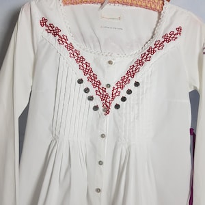 May include: White long-sleeve button-up shirt with red embroidery and silver buttons. The shirt has a gathered neckline and a small label that reads "21 republic no. 5100 canning".