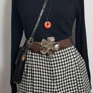 May include: A black long-sleeved shirt with a brown belt and a large silver flower buckle. The belt is worn over a black and white houndstooth skirt. A black leather purse with a gold clasp is worn on a strap over the shoulder.
