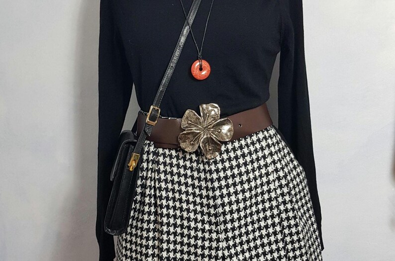 May include: A black long-sleeved shirt with a brown belt and a large silver flower buckle. The belt is worn over a black and white houndstooth skirt. A black leather purse with a gold clasp is worn on a strap over the shoulder.