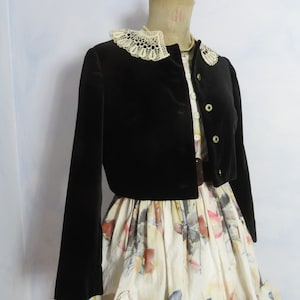 Könnte beinhalten: Eine schwarze Samtjacke mit einem weißen Spitzenkragen. Die Jacke wird über einem weißen Kleid mit Blumendruck getragen.
