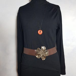 May include: A black turtleneck sweater with a brown leather belt with a large silver flower buckle. A necklace with a small orange pendant hangs from the neck of the sweater.