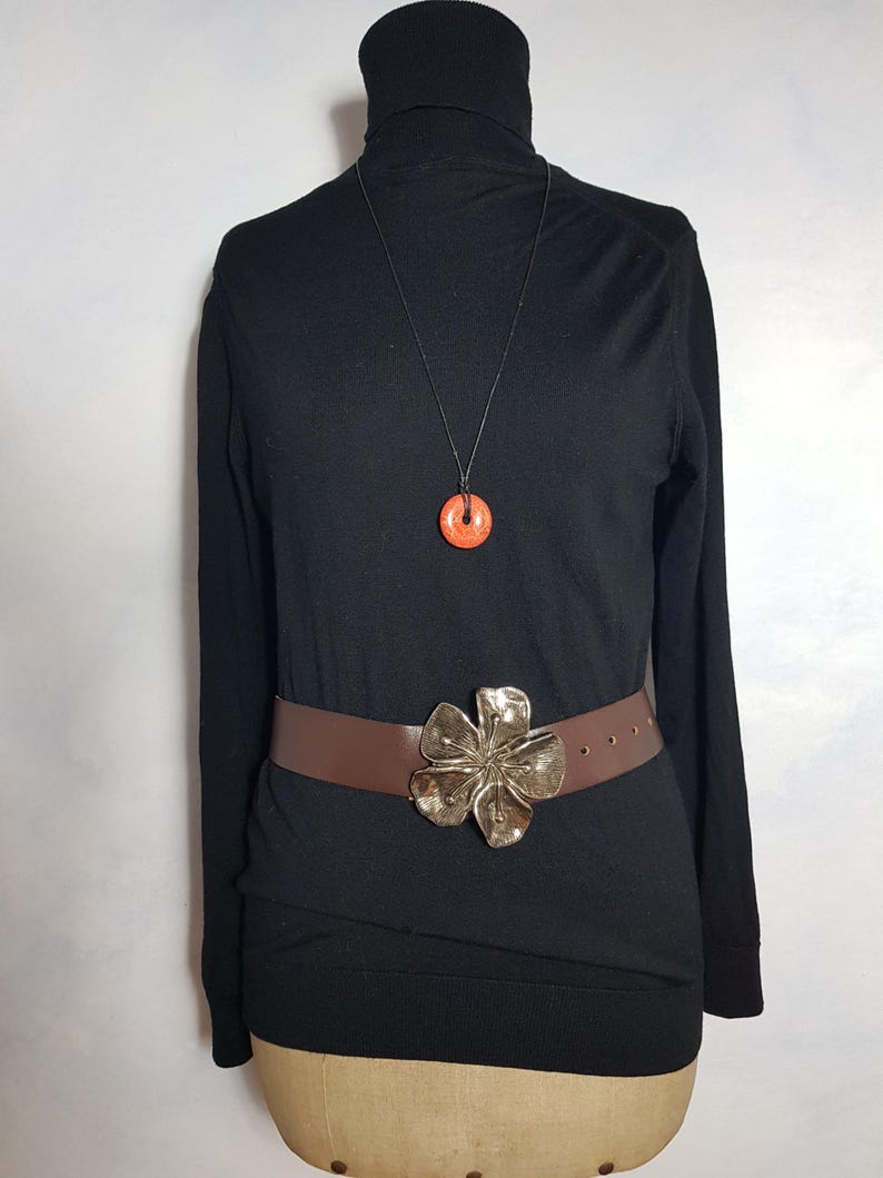 May include: A black turtleneck sweater with a brown leather belt with a large silver flower buckle. A necklace with a small orange pendant hangs from the neck of the sweater.