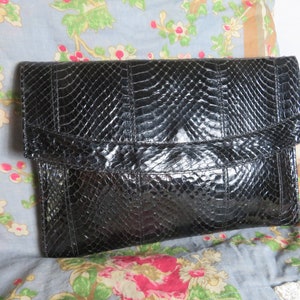 Può includere: Una pochette nera in pelle di serpente con chiusura a lembo. La pochette ha un motivo testurizzato e goffrato.