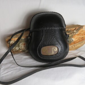 May include: A black leather bag with a long strap. The bag has a decorative metal piece on the front with a design.