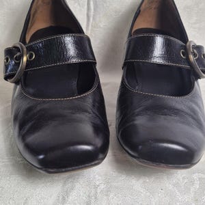 May include: A pair of black leather Mary Jane style shoes with square toes. Each shoe has a strap across the top with a large, round, bronze-colored buckle. The shoes are on a white patterned surface.
