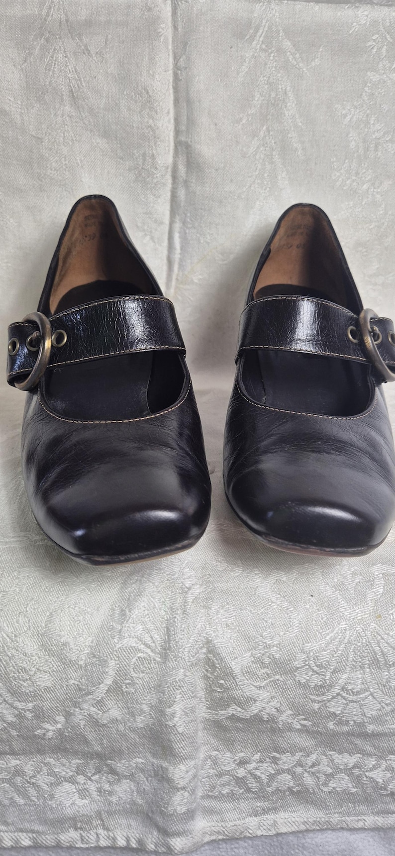 May include: A pair of black leather Mary Jane style shoes with square toes. Each shoe has a strap across the top with a large, round, bronze-colored buckle. The shoes are on a white patterned surface.