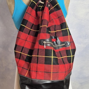 May include: Red and black plaid drawstring backpack with a black leather bottom and a zippered pocket. The bag has a black leather handle and a drawstring closure. The bag is displayed on a mannequin.