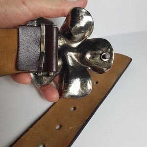 May include: A brown leather belt with a large silver flower buckle. The buckle is detailed and has a textured surface.