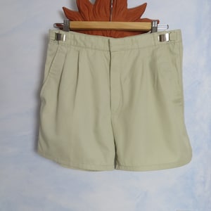 May include: A pair of light beige shorts with pleats and a button fastening. The shorts are hanging on a wooden hanger with a decorative sun-shaped design.