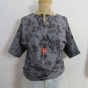 Puede incluir: Una blusa gris de manga corta con estampado floral negro y un collar con un colgante rojo.