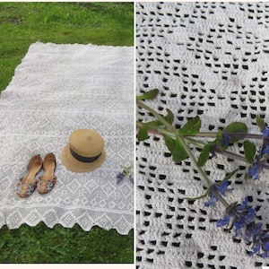 Può includere: Tovaglia da picnic in pizzo bianco con un motivo delicato, stesa sull'erba verde. Un cappello di paglia e sandali marroni sono posizionati sulla tovaglia. Un primo piano della tovaglia mostra un rametto di fiori blu.