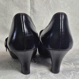 May include: Pair of black leather pumps with a low heel. The shoes have a rounded toe and a small buckle detail on the side. The heels are approximately 2.5 inches (6.35 cm) high.