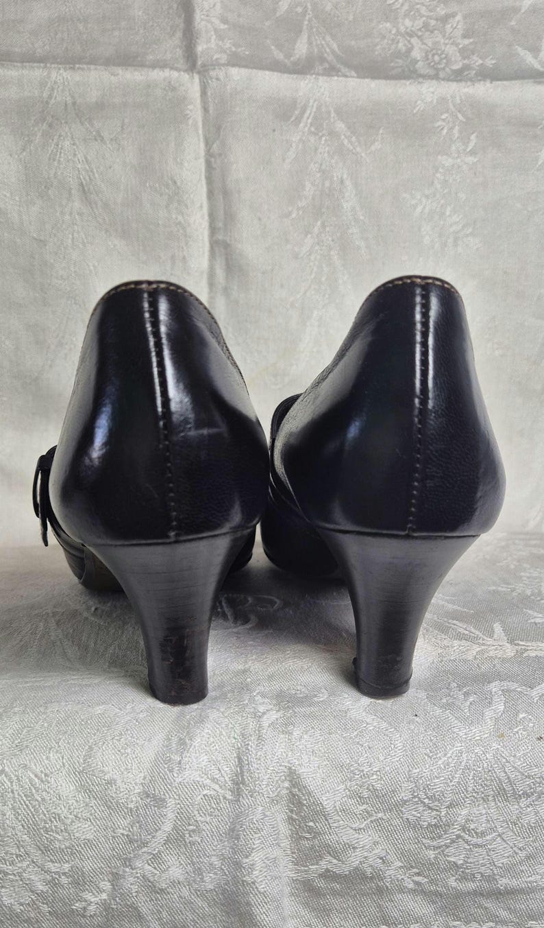May include: Pair of black leather pumps with a low heel. The shoes have a rounded toe and a small buckle detail on the side. The heels are approximately 2.5 inches (6.35 cm) high.