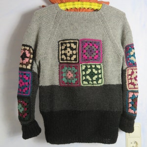Könnte beinhalten: Ein grauer und schwarzer Strickpullover mit bunten gehäkelten Quadraten an den Ärmeln und vorne. Der Pullover hat einen Rundhalsausschnitt und lange Ärmel.