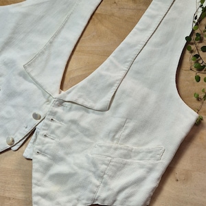 Könnte beinhalten: Eine cremefarbene, strukturierte Weste mit Knopfleiste und zwei kleinen Taschen. Die Weste ist aus einem gewebten Stoff gefertigt und liegt auf einer Holzoberfläche. Die West hat ein klassisches, maßgeschneidertes Design.