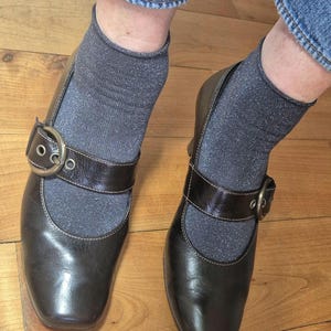 May include: A pair of dark brown leather Mary Jane style shoes with a square toe and a buckle closure. The shoes are worn with gray socks and blue jeans. The shoes are on a wooden floor.