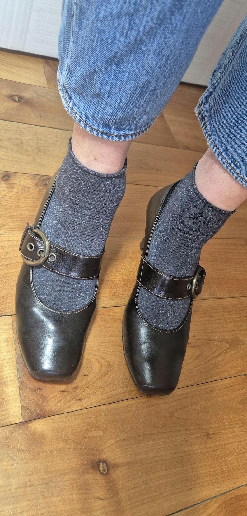 May include: A pair of dark brown leather Mary Jane style shoes with a square toe and a buckle closure. The shoes are worn with gray socks and blue jeans. The shoes are on a wooden floor.