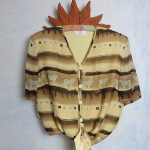 Könnte beinhalten: Eine braune und beige Seidenbluse mit einem Bindeband vorne und einem V-Ausschnitt. Die Bluse hat ein Muster aus kleinen Quadraten und Tier-Silhouetten. Die Bluse hat ein Etikett mit der Aufschrift "VAN by Valentina".