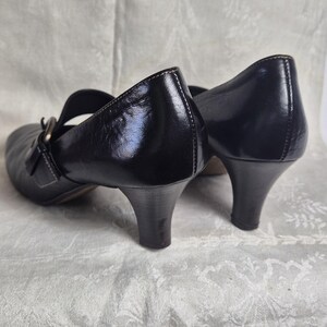 May include: Black leather Mary Jane style shoes with a low heel. The shoes have a strap across the top of the foot and a buckle closure. The heels are approximately 2 inches high.