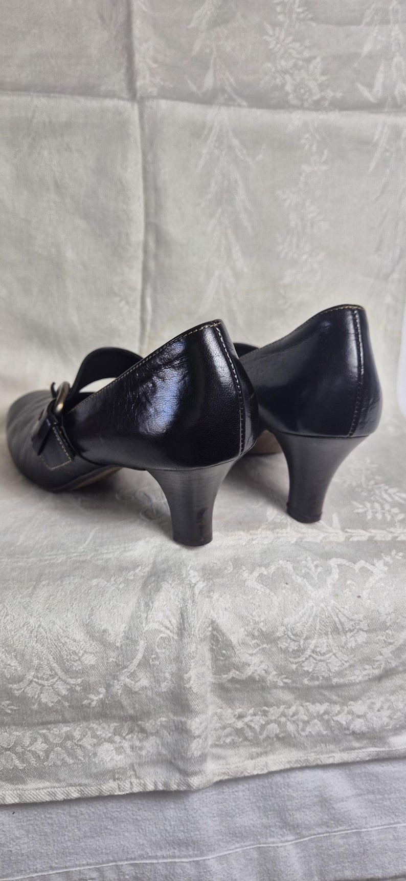 May include: Black leather Mary Jane style shoes with a low heel. The shoes have a strap across the top of the foot and a buckle closure. The heels are approximately 2 inches high.