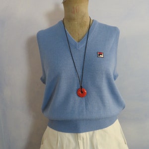 May include: A light blue sleeveless sweater with a v-neck and a small logo on the right side. The sweater is worn over a white skirt and a red pendant necklace.