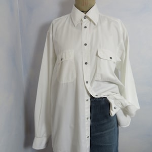 May include: A white long-sleeved button-down shirt with two flap pockets and a button-down collar. The shirt is on a mannequin and is unbuttoned, revealing a pair of blue jeans underneath.