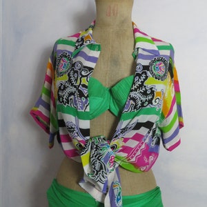 May include: A colourful striped shirt with a paisley pattern tied around a green bikini top. The shirt has a white background with stripes of pink, purple, green, yellow, and black. The paisley pattern is in black, white, and shades of green.