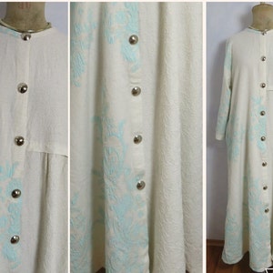 Może przedstawiać: Długa, biała, zapinana na guziki sukienka z jasnoniebieskim wzorem kwiatowym. Sukienka ma kołnierz i długie rękawy.