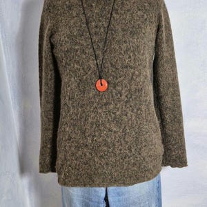 Könnte beinhalten: Ein brauner Pullover mit rundem Ausschnitt und langen Ärmeln. Der Pullover wird mit hellblauen Jeans kombiniert. Eine Halskette mit rotem Anhänger hängt um den Hals.