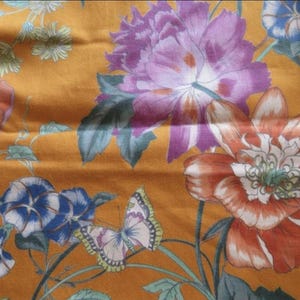 Puede incluir: Primer plano de una tela con un estampado floral. El diseño presenta grandes flores en tonos naranja, morado y blanco, junto con campanillas azules y una mariposa, todo sobre un fondo amarillo dorado.