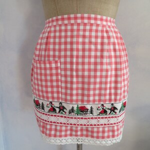 May include: A pink and white gingham apron with a white lace trim and a pocket. The apron has a band of embroidered figures and trees along the bottom.
