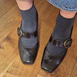 May include: A pair of dark brown Mary Jane style shoes with a square toe and a strap with a buckle. The shoes are worn with gray socks and denim jeans. The shoes are on a wooden floor.