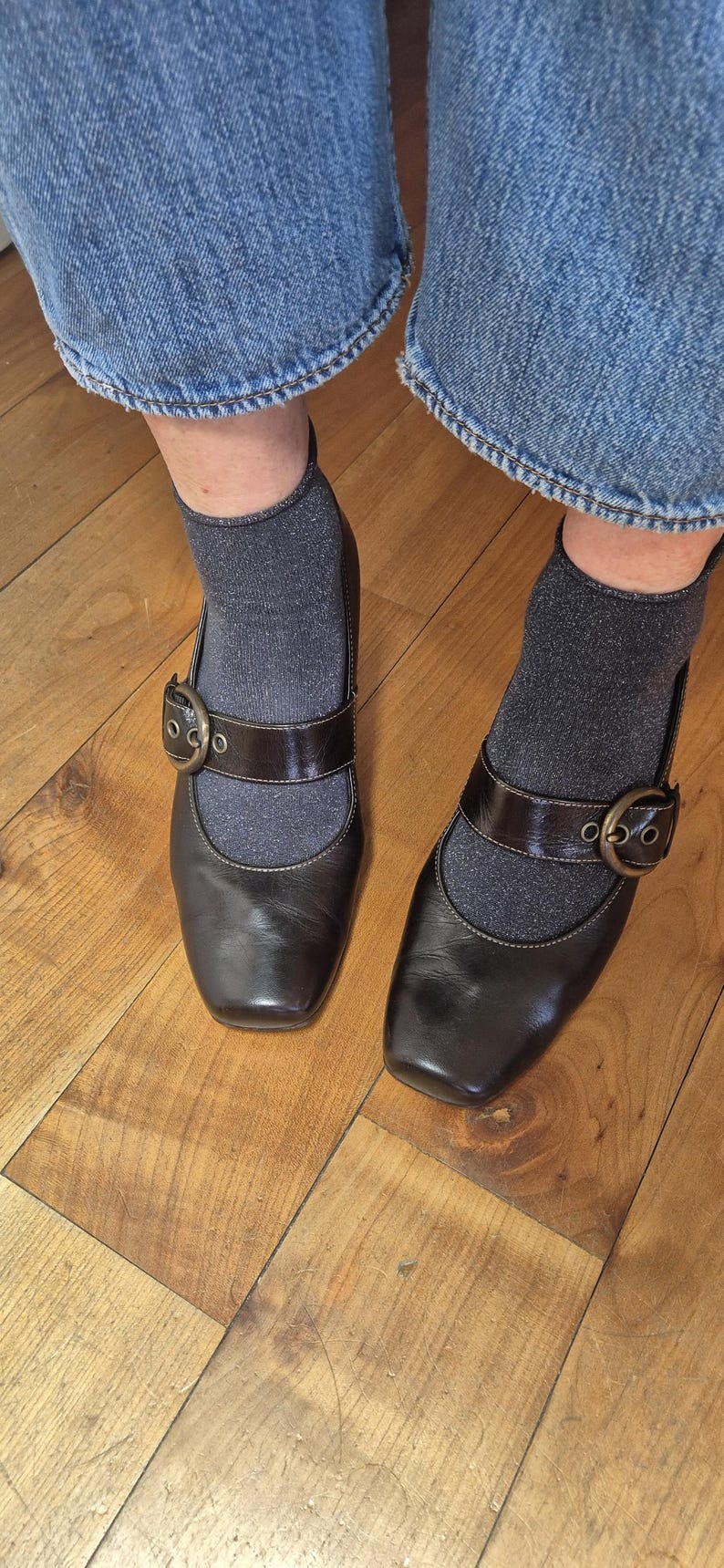 May include: A pair of dark brown Mary Jane style shoes with a square toe and a strap with a buckle. The shoes are worn with gray socks and denim jeans. The shoes are on a wooden floor.