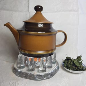 May include: A brown ceramic teapot with a lid and a glass stand with clear glass cylinders. The teapot is sitting on the stand. There is a small white dish with dried green herbs next to the teapot.