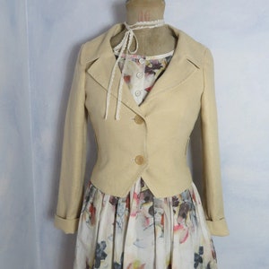 Könnte beinhalten: Eine beige, kurze Jacke mit Kragen und drei Knöpfen. Die Jacke befindet sich auf einem Mannequin, das ein weißes Kleid mit Blumendruck trägt.