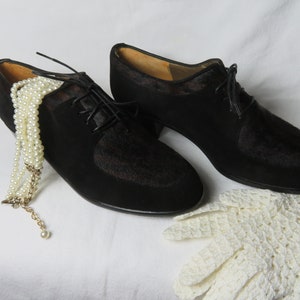 May include: A pair of black leather lace-up shoes with a low heel. The shoes are on a white surface with a pearl necklace and a white crocheted glove.
