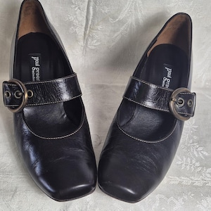 May include: A pair of black leather Mary Jane style shoes with a square toe and a buckle closure. The shoes have a small heel and are displayed on a patterned surface. The brand name "paul green" is visible on the insole.